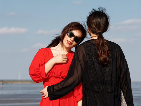 Beautiful Chinese girls dressed in red dress and black dress hugging together on sunny beach. Best young girl friends enjoy carefree time in summer, Emotions, people, beauty and lifestyle concept.