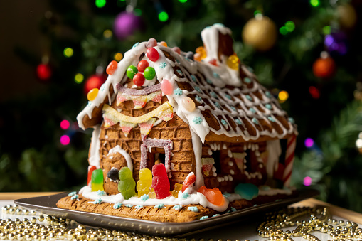Homemade gingerbread house decorated with icing, sweets , and jelly candies with blurred Christmas tree on background. Family Christmas