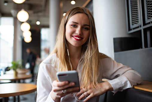 Cheerful young blond lady in blouse messaging on mobile phone while relaxing in modern cafe and toothy smiling at camera