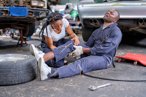 Mechanical guy at car repair shop got accident sit on the floor mechanical girl first aid check his broken leg painful and emergency call for help