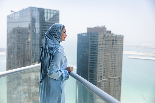 Mature middle eastern woman observes Dubai from her home. She is amazed by the view. She is wearing traditional blue dress. Focus on foreground.