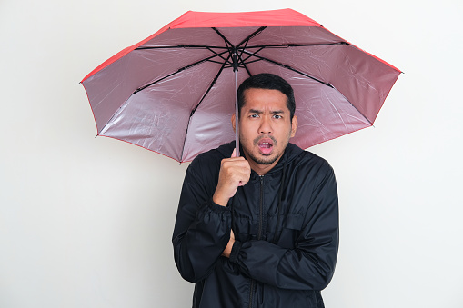 Adult Asian man wearing jacket and using an umbrella with worried expression