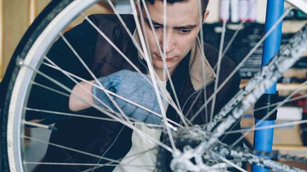 istock Young man experienced serviceman is fixing bike wheel using wrench and tools. Small cozy workshop interior with wooden walls and ladder, spare parts and equipment are visible. 1437023144