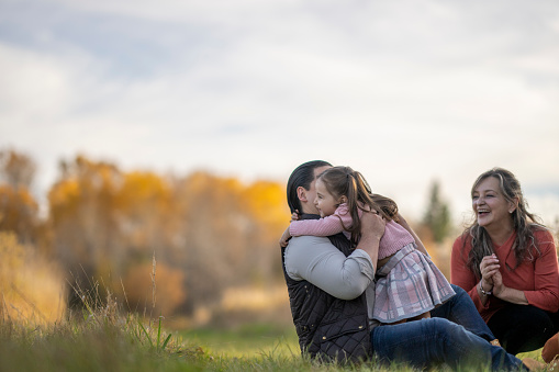 A small family of three consisting of a Father, Grandmother and Granddaughter, spend time outside on a sunny fall day.  They are each dressed comfortably in layers as they sit in the grass talking and enjoying the crisp fresh air.