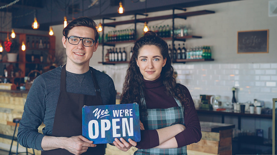 Cheerful attractive young people small coffee-house owners are holding we are open sign while standing inside modern coffee shop. Opening new business and people concept.