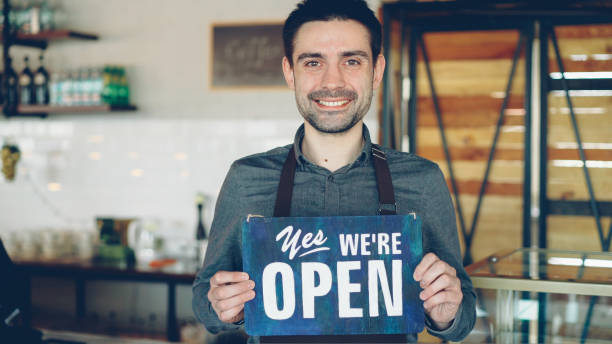 istock Cheerful handsome man small coffee-house owner is holding We Are Open sign while standing inside coffee shop. Opening new business and people concept. 1437035952