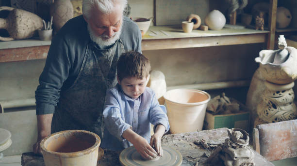 istock Young cute boy is making clay figure on throwing wheel while his grandfather experienced sculptor is standing near worktable and watching carefully. Family tradition concept. 1437037857