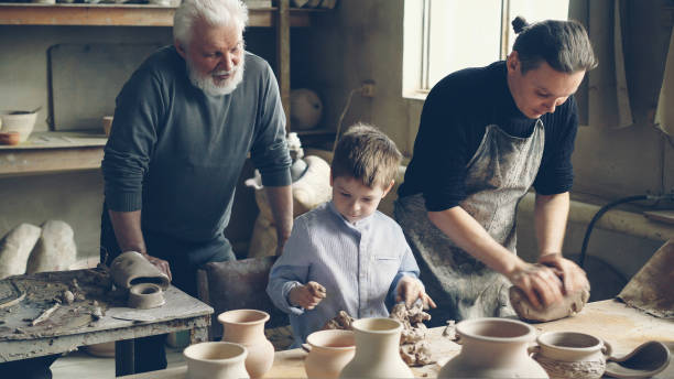istock Professional potter is kneading clay on worktable in home studio while his son is helping him and his elderly father watching them from behind. Small family business concept. 1437038238