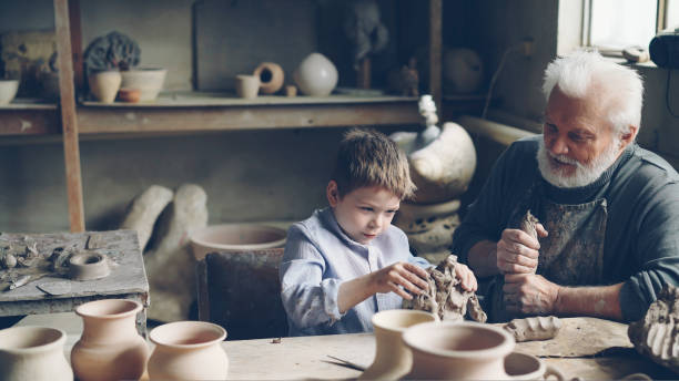 istock Cute boy is having fun playing with clay in his grandfather's pottery studio while elderly man is kneading clay preparing to mold utensils. Family spending time together concept. 1437038793
