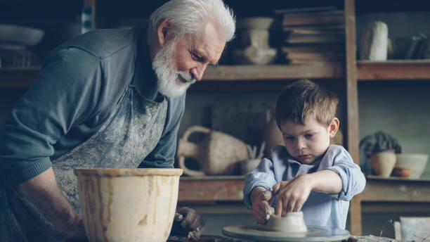 istock Concentrated young boy is molding clay into ceramic pot on spinning throwing wheel and his experienced grandfather is talking to him. Pottery and family tradition concept. 1437038918