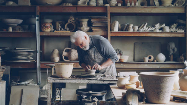 istock Experienced ceramist is creating utensils from clay on potter's wheel in workshop. Producing eathenware, handmade utensils and professional pottery concept. 1437039873