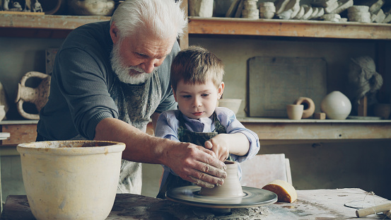 Concentrated little boy is learning to work with clay on professional throwing-wheel in pottery class in traditional workshop. His teacher senior experienced man is helping him.
