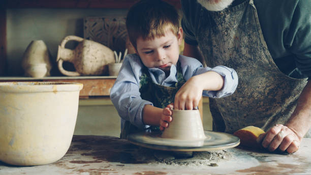 istock Curious boy is learning pottery from his experienced grandfather in small home studio. Child is forming clay to make pot on potter's wheel, his granddad is helping him. 1437041450