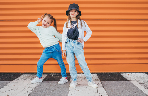 Dos hermanas pequeñas vestidas con ropa de estilo moda infantil posando ante la cámara en la carretera peatonal de asfalto de la ciudad cerca del fondo naranja de la pared. Feliz imagen conceptual de la infancia moderna. photo