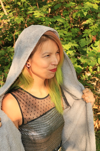 An Indigenous woman in a forest smiling on a sunny day. She is wearing an open gray hooded sweater, and a silver and black dress with earrings and a nose ring.