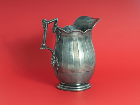Antique pewter water pitcher centered on red background. Highly decorated pitcher etched and embossed with ornate handle.