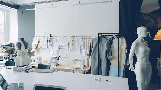 Light clothing design studio with large tailor's desk, mannequins, numerous sketches pinned on wall, sewing machines, tailoring items and half-finished garments on rails.