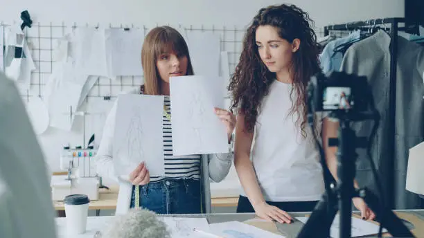 Photo of Creative bloggers tailors recording video about new sketches. Geirls are greeting followers, showing drawings and talking emotionally. Camera and tailoring items are visible.