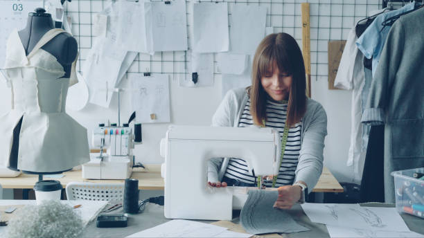 istock Young attractive dressmaker is working on sewing machine and looking at women's garment sketches in her studio at table. Modern equipment and fashionable garments in background. 1437050764