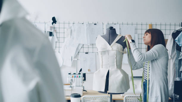 istock Pretty young woman is adjusting clothes on tailoring dummy with pins while working at new women's clothes in light tailor's shop. Creating new garment concept. 1437051909