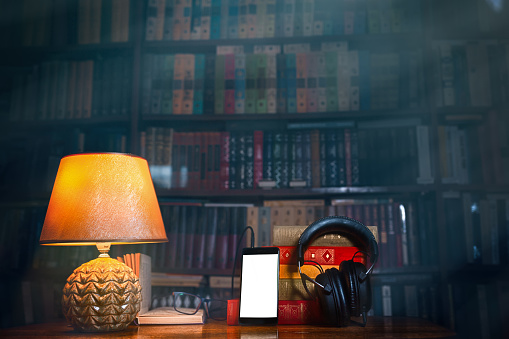 On a wooden table next to the lamp are headphones, a phone, a stack of books and glasses. The concept of modern technology and audiobooks. Tint and copy space.