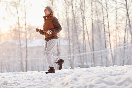 Side view portrait of sporty mature woman running in winter forest and smiling, copy space