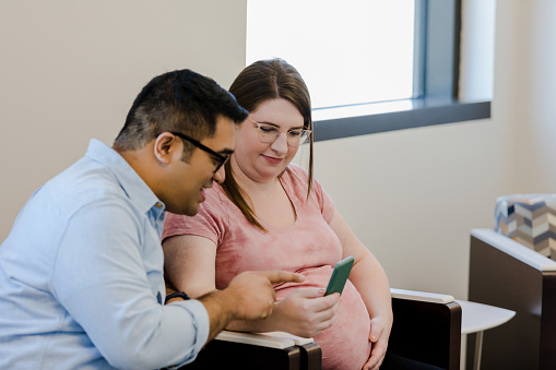 In the lobby of the clinic, the young adult expectant couple views ultrasound images of their baby on the smart phone.