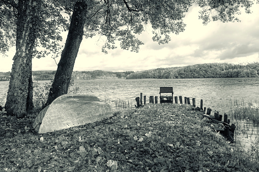Autumn lake scenery with a boat and a chair at shore, black and white processed