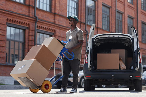 Full length portrait of smiling delivery worker unloading boxes outdoors in sunlight, copy space