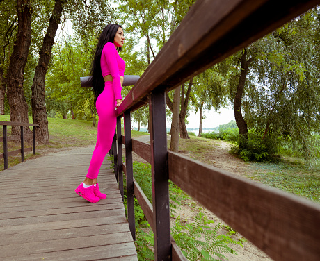 A sports woman in a pink suit stands on a wooden bridge. Portrait of a woman with dark hair. The woman is standing. woman looking