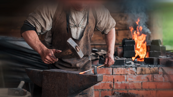 Hands of a man with a hammer on the background of an authentic retro forge with bright fire. Added motion blur effect for cinematic look.