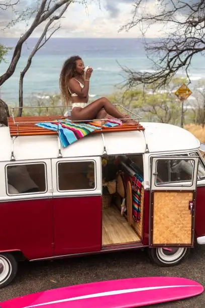 A cool summer shot of a black woman in a bikini sitting on a Volkswagen van on the beach in Hawaii