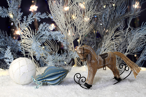 Wooden toy rocking horse stands on artificial snow and New Year toys lie next to it and artificial blue and white trees with festive lights stand behind them against a dark blue background. Closeup. Night studio shooting..