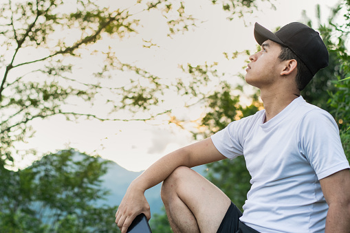 young latin man, sitting on top of a mountain looking up to the sky, meditating outdoors in the middle of the forest. brown guy with sunset in the background, he has a black cap and white shirt.