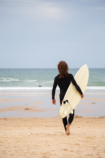 Back view of young man in black wetsuit walking towards sea with surfboard on daytime