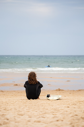 Back view of relaxed sportsman in black wetsuit relaxing on sandy beach and contemplating seascape on daytime