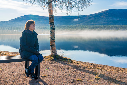 Woman sitting on a bench at a beach watching the nice view with mountain and river, Gällivare, Swedish Lapland, Sweden