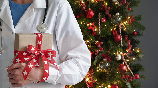 Concept for christmas and new year medical banner.Female doctor in white coat,stethoscope holds gift box with red bow in her hands against background beautifully decorated Christmas tree.Copy space.