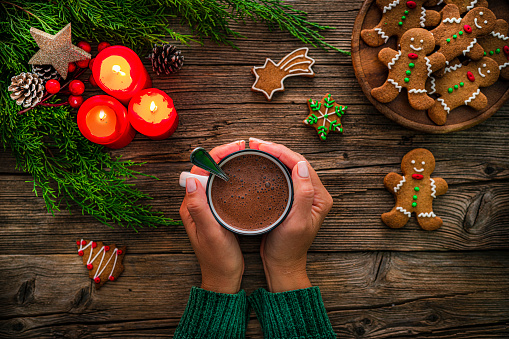 Close up view of female hands holding hot chocolate mug shot from above on rustic wooden Christmas table. Homemade Christmas cookies, burning candles and Christmas decoration complete the composition. High resolution 42Mp studio digital capture taken with Sony A7rII and Zeiss Batis 40mm F2.0 CF lens
