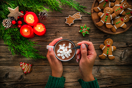 Close up view of female hands holding hot chocolate mug with marshmallows shot from above on rustic wooden Christmas table. Homemade Christmas cookies, burning candles and Christmas decoration complete the composition. High resolution 42Mp studio digital capture taken with Sony A7rII and Zeiss Batis 40mm F2.0 CF lens