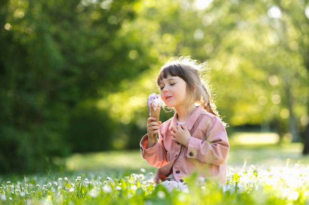 счастливый ребенок ест мороженое, сидя на траве в парковом портрете. счастливое детство. - baby spring child grass стоковые фото и изображения
