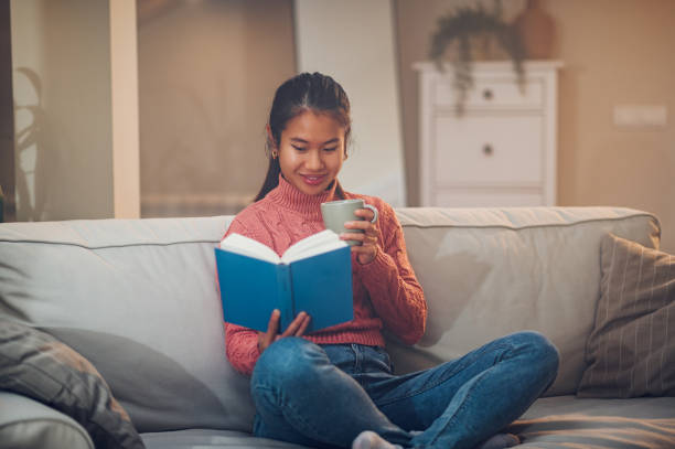 istock Asian woman siting on the couch at home while reading a book and drinking coffee 1437100918