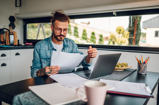 Smiling young tattooed man in jeans shirt working on a laptop in modern kitchen and holding work documents. Concept of young people working from home.