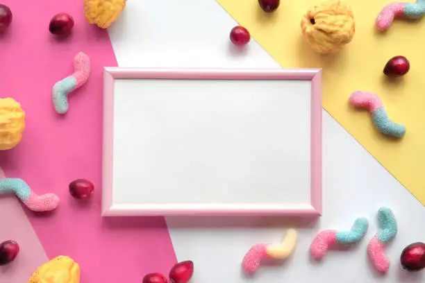 Blank pink white frame on layered colored paper. Assorted sweets, confectionery and cranberry berry around empty frame with copy-space, place for text. Flat lay, top view. Colorful greeting design.