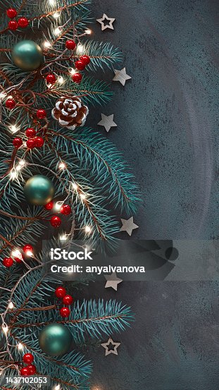 istock Christmas vertical background for social media stories. Dark flat lay on fir twigs, red berries, cones and light garland. 1437102053