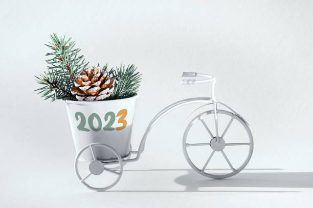 2023 caption, greeting on white decorative bike. Bicycle with pine cone and fir twigs in decorative basket. Happy New Year greeting design. Off white background, sunlight, long shadows. stock photo