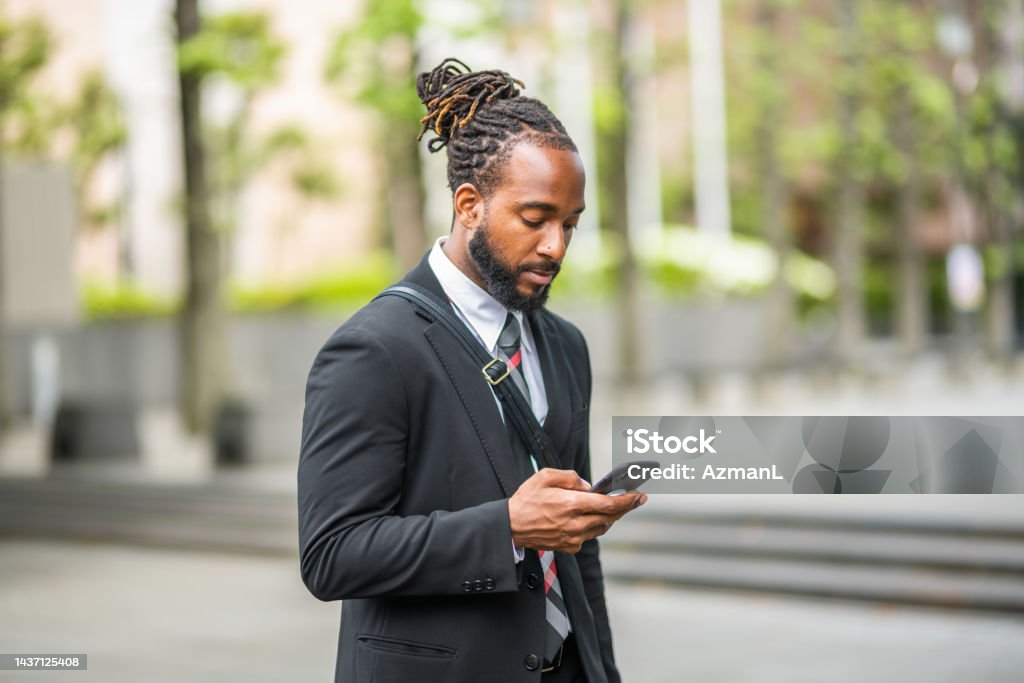 Black Male Lawyer Texting And Walking Outdoors Waist up of a handsome black male lawyer working on his smart phone while walking towards his office. He is outdoors in the city wearing a black suit and a tie. Lawyer Stock Photo