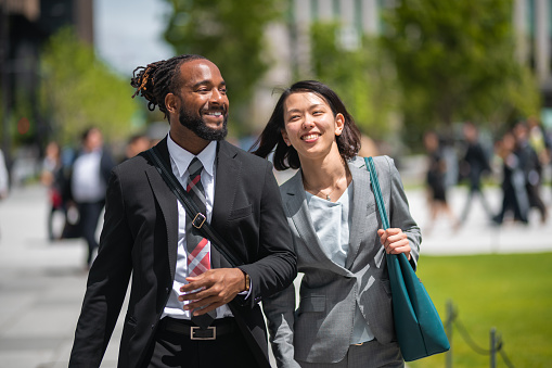 African American male accountant and Chinese female manager going to work. They are smiling and talking about their holidays. They are outdoors in the city. Defocused city park and people in the background.
