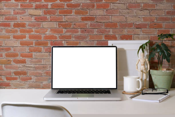 istock Front view laptop computer with blank screen, picture frame, glasses and houseplant on white table against brick wall. 1437136922