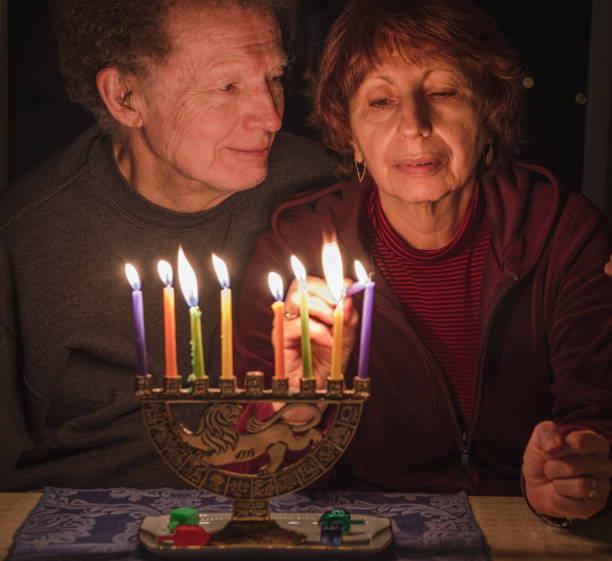 la donna anziana accende le candele a casa per celebrare la festa ebraica di hanukkah; uomo che la guarda affettuosamente - adult senior adult traditional culture usa foto e immagini stock